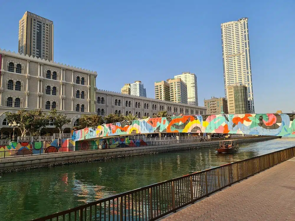 Colorful mural-covered bridge over a canal with tall Al Qasba city buildings in the background