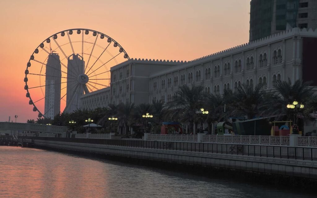Ferris wheel beside a waterfront building at sunset, with palm trees and glowing lamps