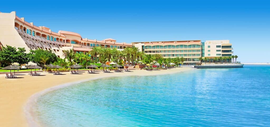 Luxury Al Raha Beach beachfront resort with palm trees, lounge chairs, and a curved sandy bay under a clear blue sky
