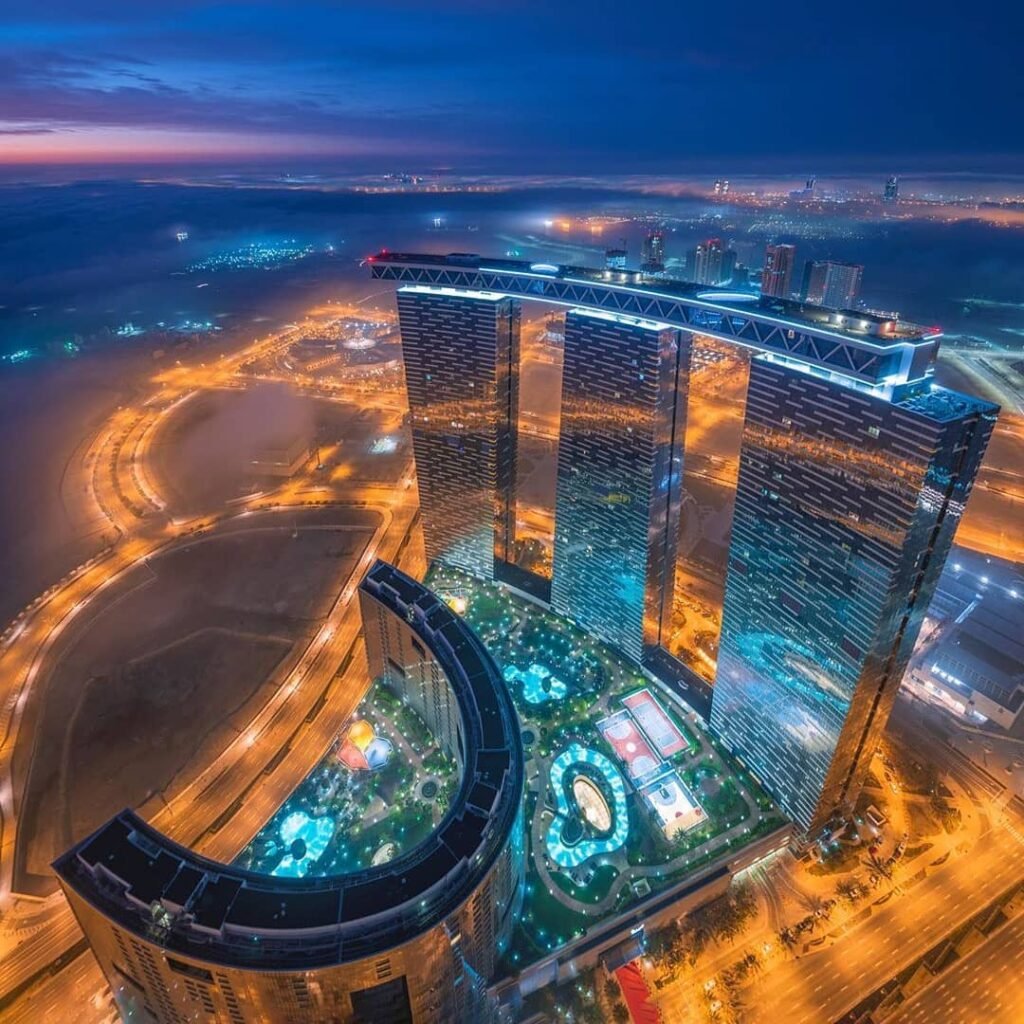 Aerial view of Al Reem Island illuminated skyscrapers with a rooftop skybridge and glowing pools at dawn