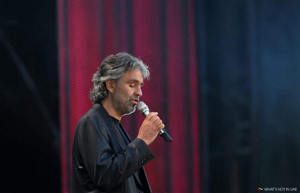 Andrea Bocelli speaking into a microphone onstage against a red and blue lit background