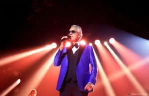 An older man in a blue suit and sunglasses sings into a microphone under bright stage lights