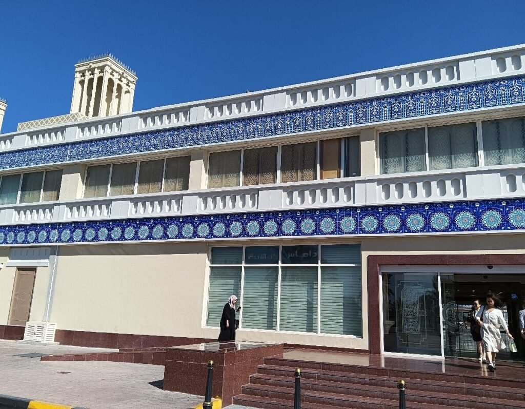 Ornate beige building with blue tile bands and a tower, viewed under a clear sky, with people at the entrance