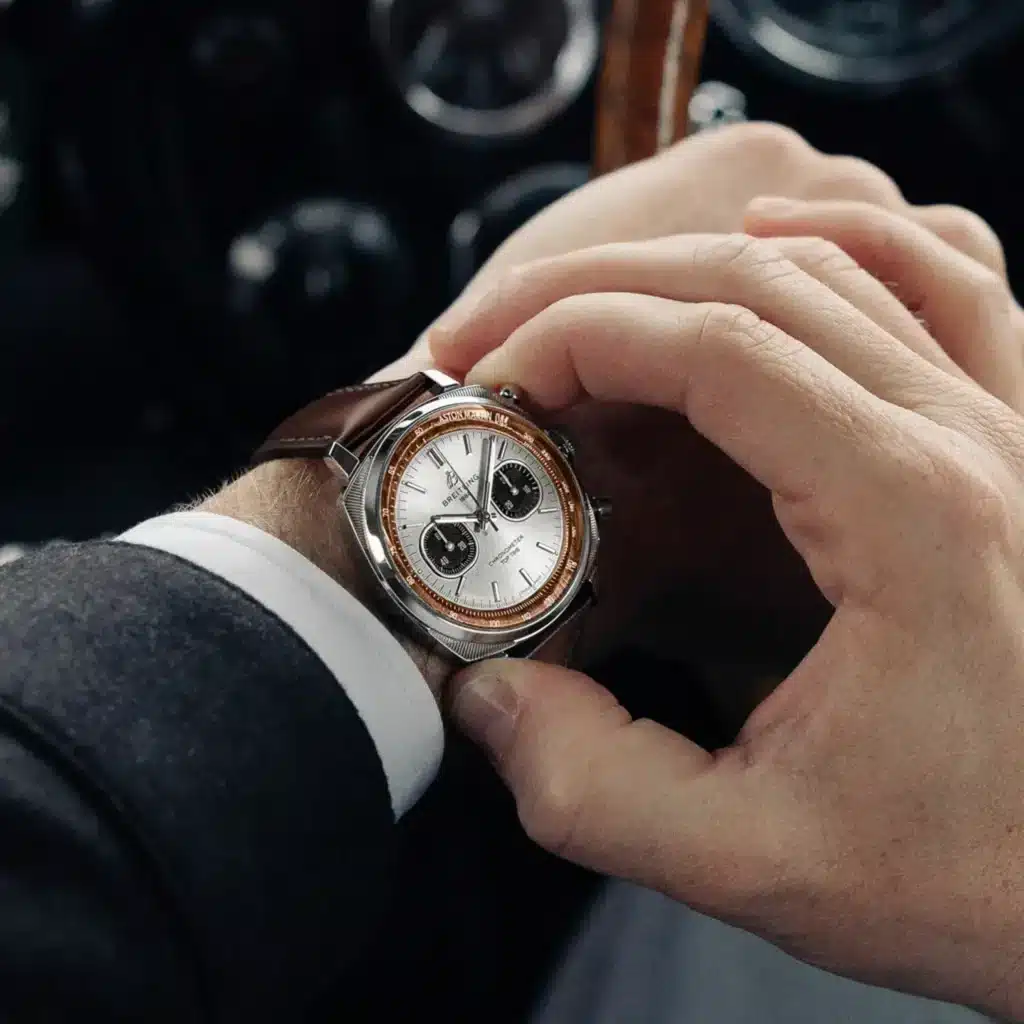 Hands adjusting a Breitling and Aston Martin DB5 chronograph watch with a brown leather strap on a wristuitcase