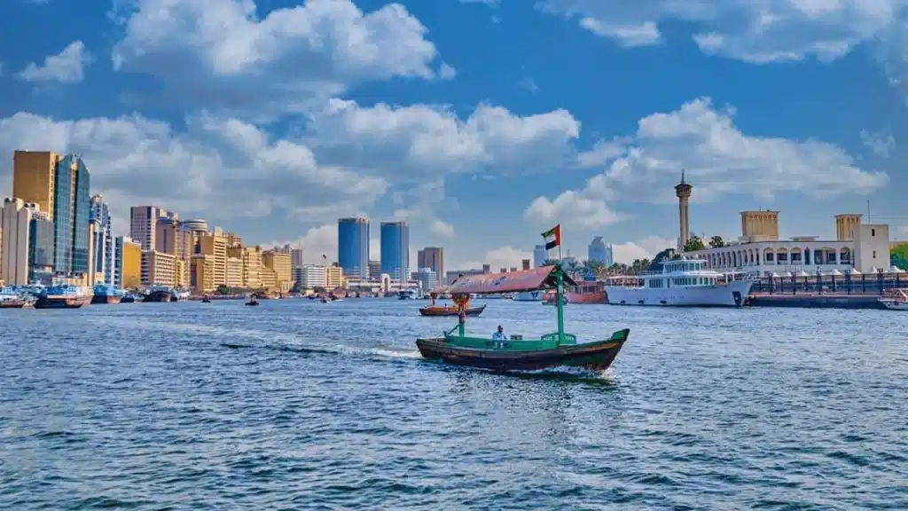 Boat crossing Bur Dubai with a city skyline, tall buildings, and a blue sky with clouds