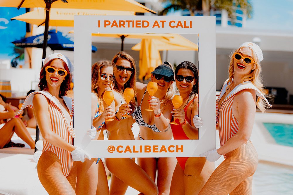 Women in striped swimsuits pose around a photo frame at a pool party, holding orange drinks and smiling