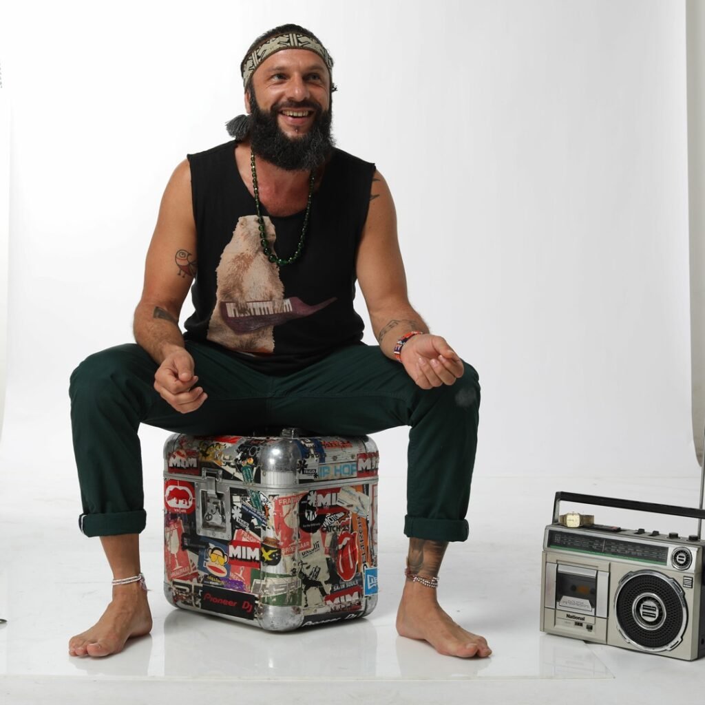 Smiling Charl Chaka sitting barefoot on a sticker-covered case beside a retro radio against a white background