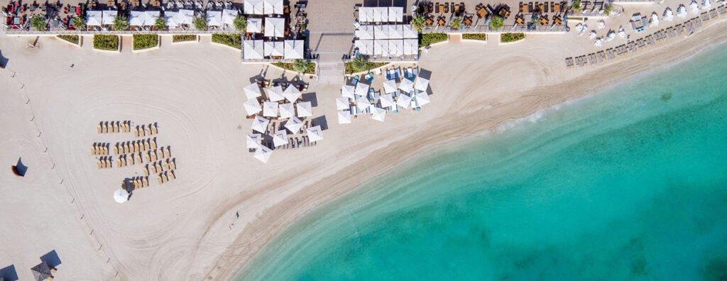 Aerial view of a beach resort with white sand, turquoise water, lounge chairs, and rows of cabanas and umbrellas
