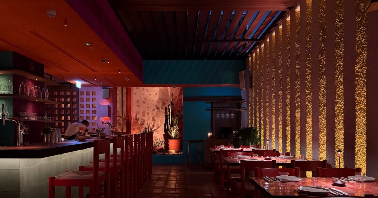 Dimly lit modern restaurant with red chairs, set tables, a bar, and a glowing textured wall at night