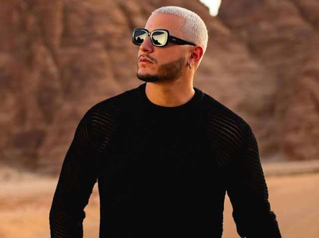 DJ Snake with bleached hair and sunglasses wearing a black shirt in a desert landscape