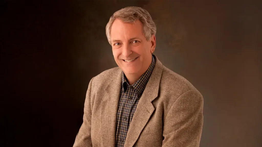 Smiling gray-haired man in a tan blazer and plaid shirt against a dark studio background