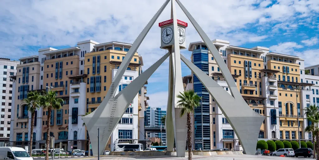 Deira clock tower framed by a white triangular sculpture in a city plaza with apartment buildings and palm trees