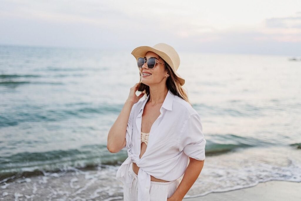 How to dress middle east heat: Woman in a sunhat and sunglasses standing on a beach by the ocean, wearing a white shirt and smiling