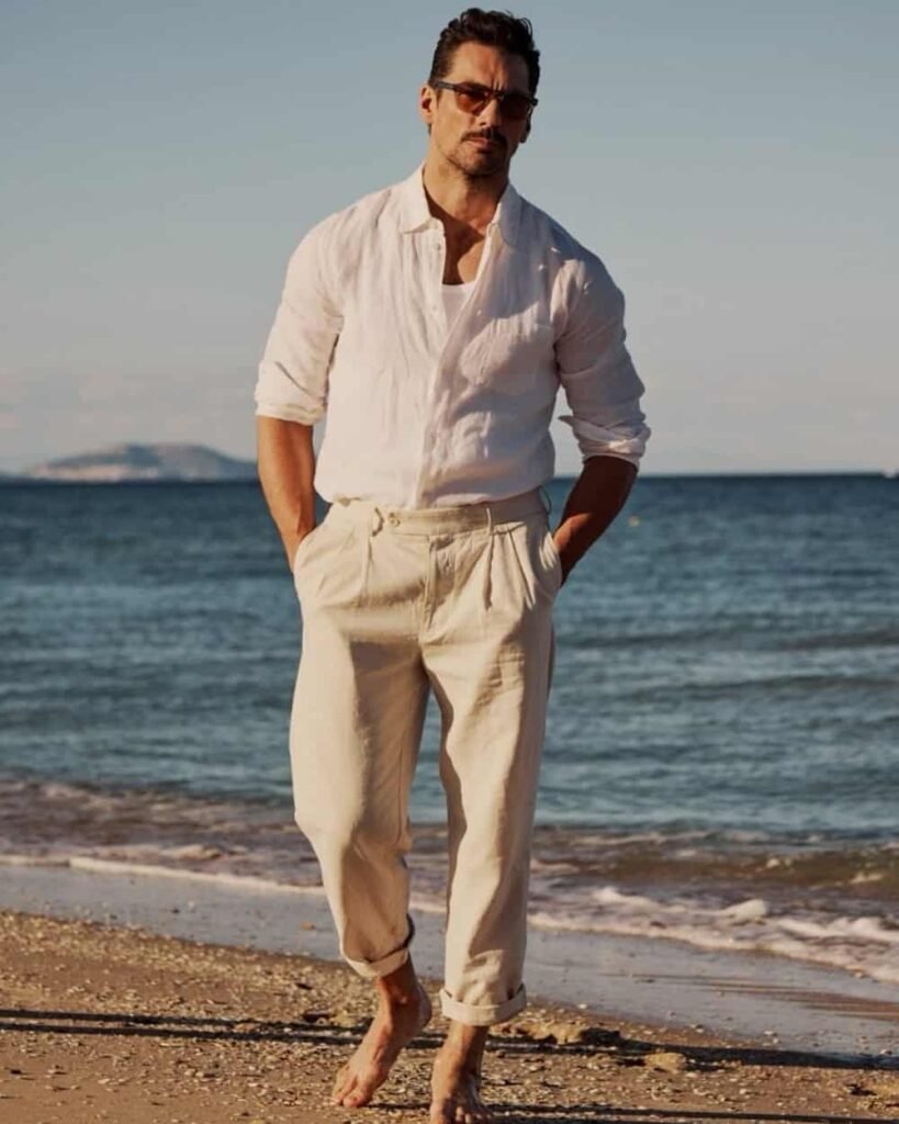 A man in sunglasses walks barefoot on a beach wearing a white shirt and beige pants