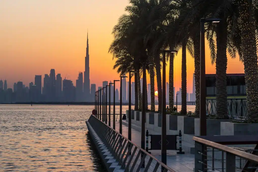 Sunset over Dubai Creek Harbour promenade with palm trees, lights, and the Dubai skyline silhouetted in the distance