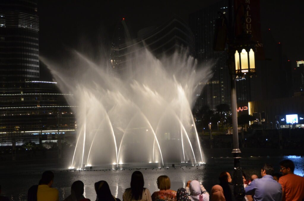 Nighttime fountain show with tall water jets lit against city skyscrapers, viewed by a crowd in the foreground
