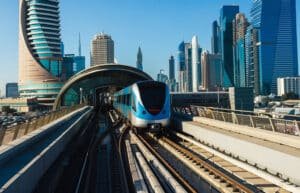 Blue metro train emerging from an elevated station amid a modern skyscraper skyline