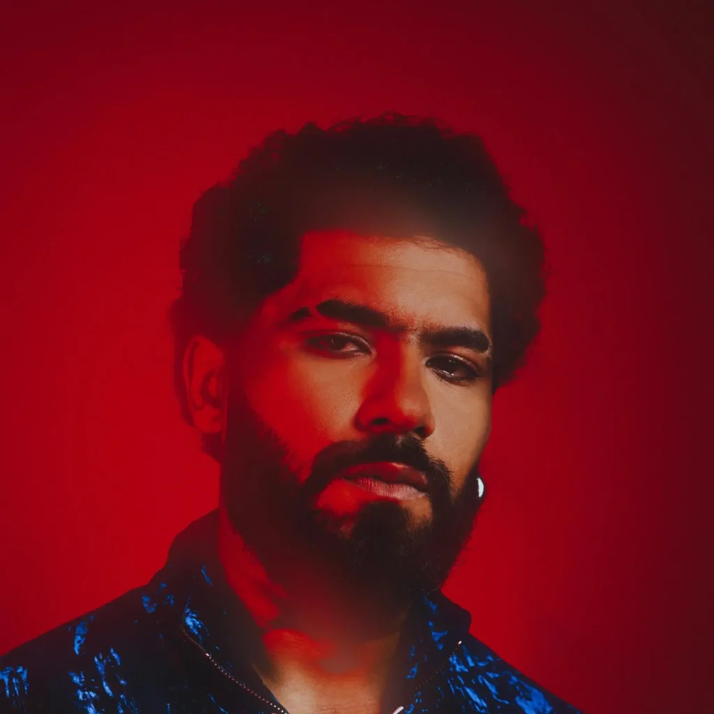 Close-up portrait of ElGrandeToto lit in red against a red background, wearing a dark blue shirt