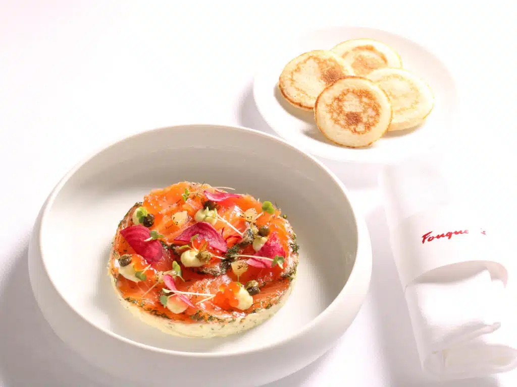 Gourmet salmon tart topped with edible flowers in a white bowl, with crisp rounds on a side plate and a folded napkin