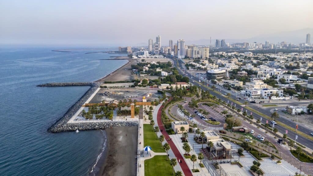 Aerial view of Fujairah City with a beach, waterfront promenade, palm trees, roads, and high-rise buildings