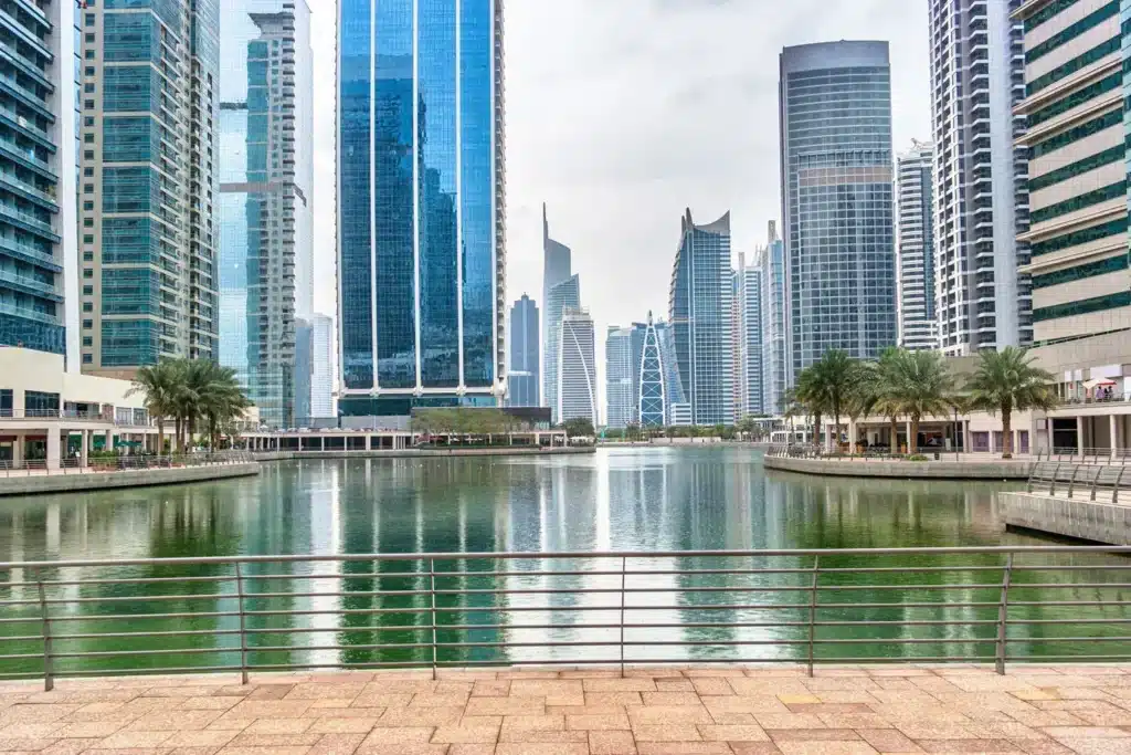 Modern waterfront JLT (Jumeirah Lakes Towers) skyline with tall glass skyscrapers reflected in a calm canal, framed by palm trees and a railing