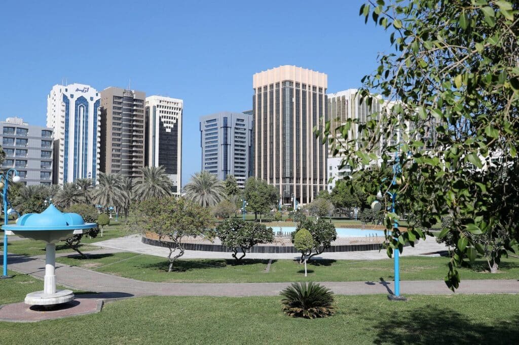 Khalidiyah Park with palm trees and a small fountain in front of modern high-rise buildings under a clear blue sky