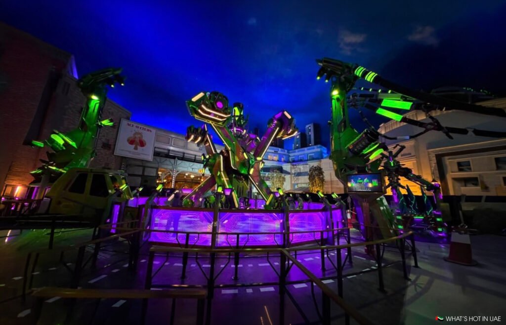 Neon-lit kryptonite collider amusement ride with giant green and purple mechanical arms in a fenced plaza at night