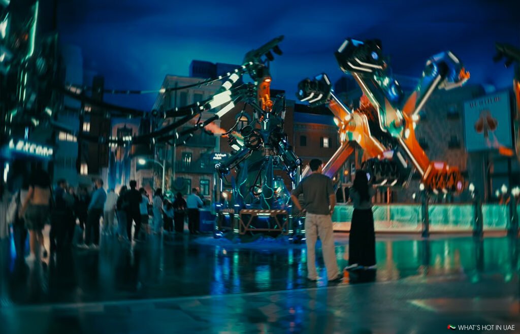 People watch a glowing mechanical kryptonite collider amusement ride with swinging arms in a nighttime plaza