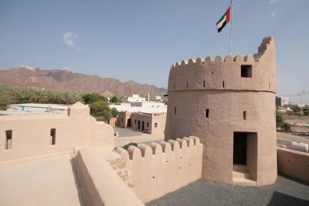 Masafi fort tower with a UAE flag, set against mountains and palm trees under a clear sky