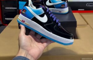 Hand holding a black, blue, and white Nike sneaker with purple laces and blue sole, with another shoe and box behind it