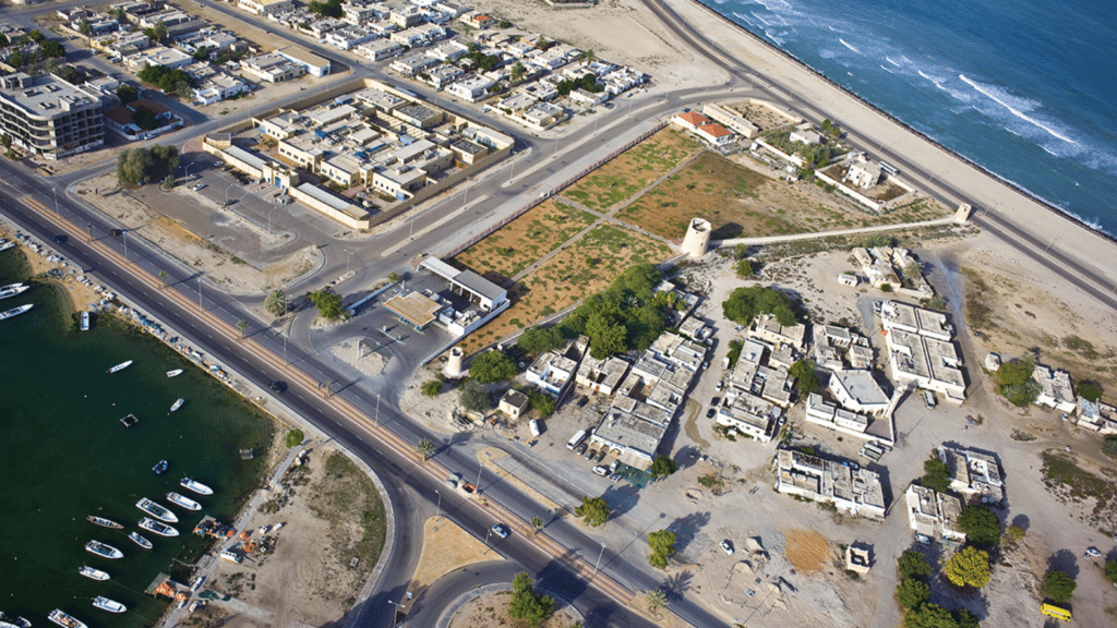 Aerial view of the Old Town in Umm Al Quwain  with roads, sandy lots, buildings, a lagoon with boats, and the ocean shoreline