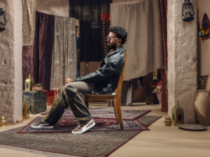 A man in a black leather jacket sits on a chair in a room with hanging patterned fabrics and rugs