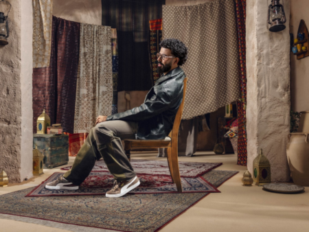 A man in a black leather jacket sits on a chair in a room with hanging patterned fabrics and rugs
