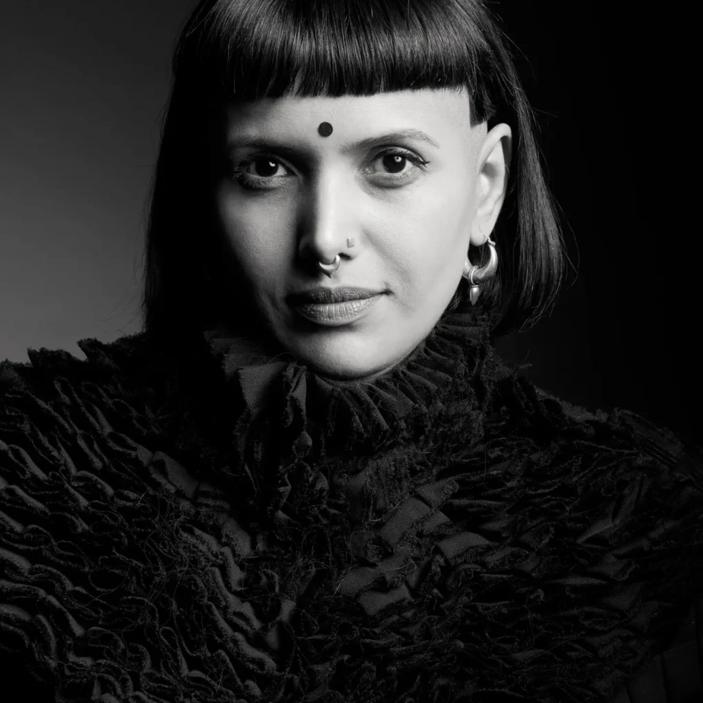 Black-and-white portrait of Pooja B with blunt bangs, facial piercings, and a textured high-collar outfit