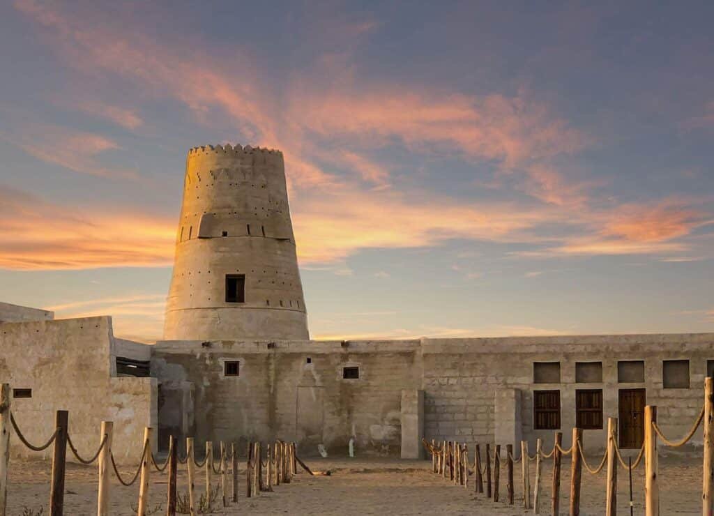 Sunset over Ras Al Khaimah stone fort with a tall round tower and pink clouds in the sky