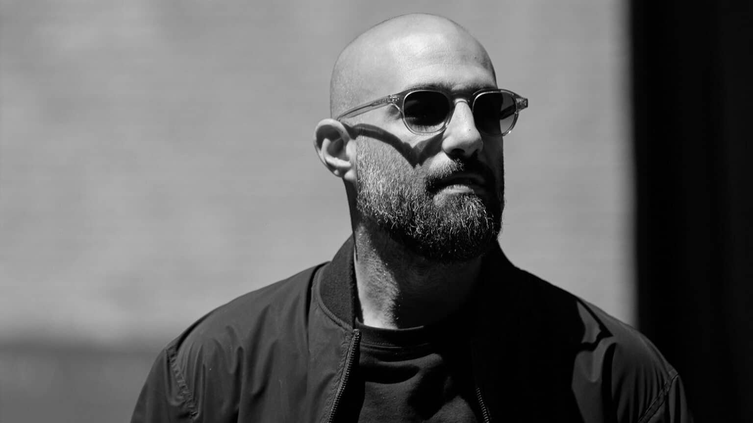 Black-and-white portrait of a bald man with a beard wearing sunglasses and a dark jacket