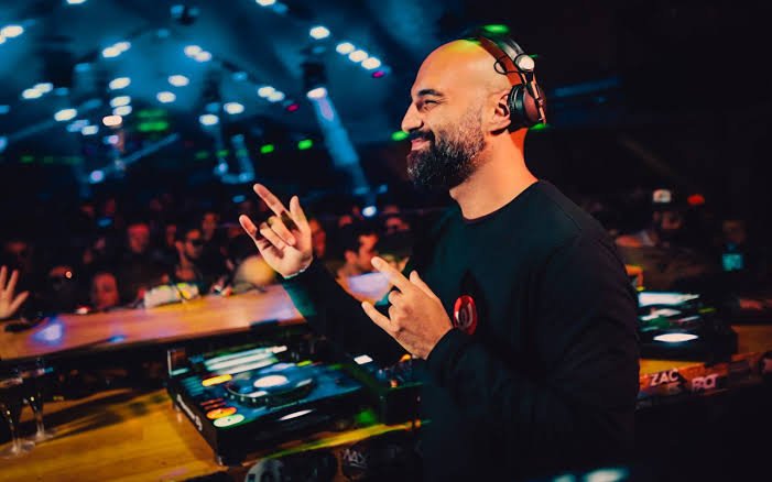 Raxon wearing headphones smiles and gestures beside a mixer in a dimly lit club