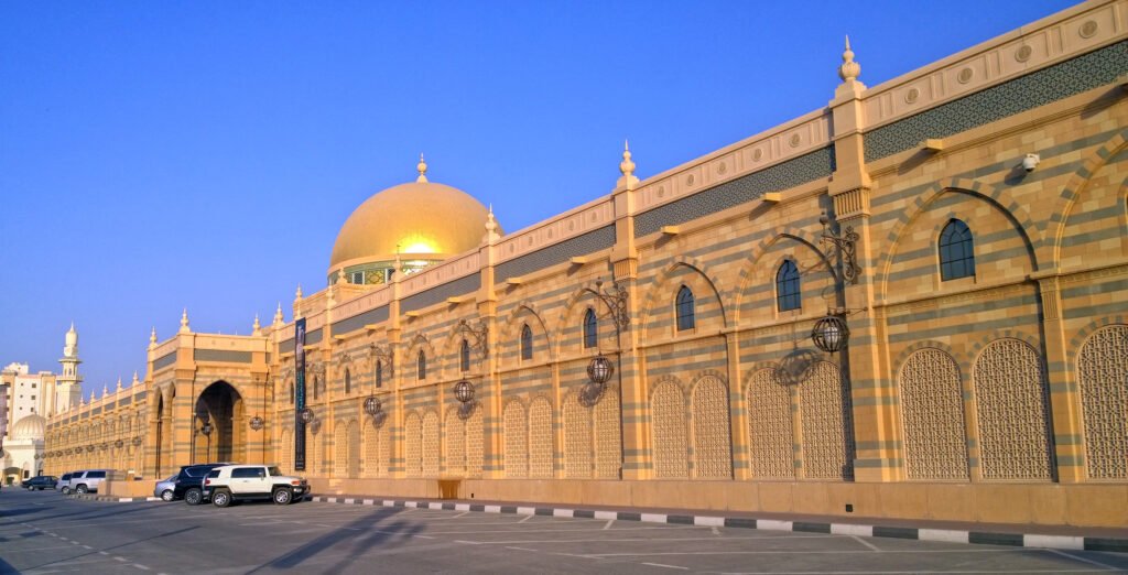 Large Sharjah mosque with a golden dome and arched windows beside an empty parking lot at dusk