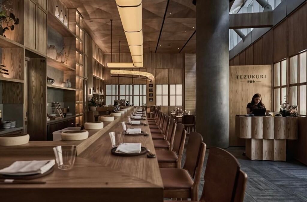 Warm wood Japanese restaurant interior with a long communal table and a reception desk labeled Tezukuri