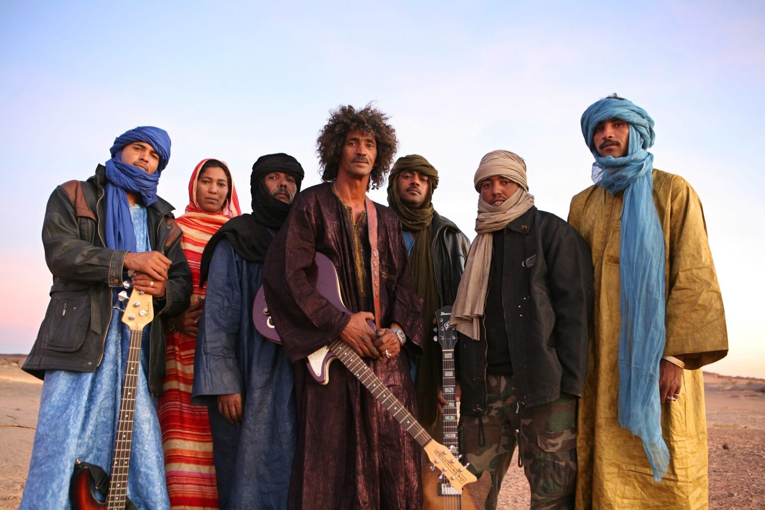 Six people in colorful robes and headwraps stand together in a desert, holding guitars and looking at the camera