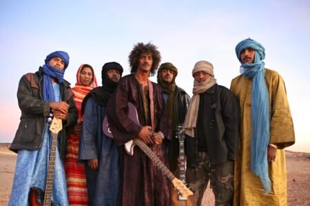 Six people in colorful robes and headwraps stand together in a desert, holding guitars and looking at the camera