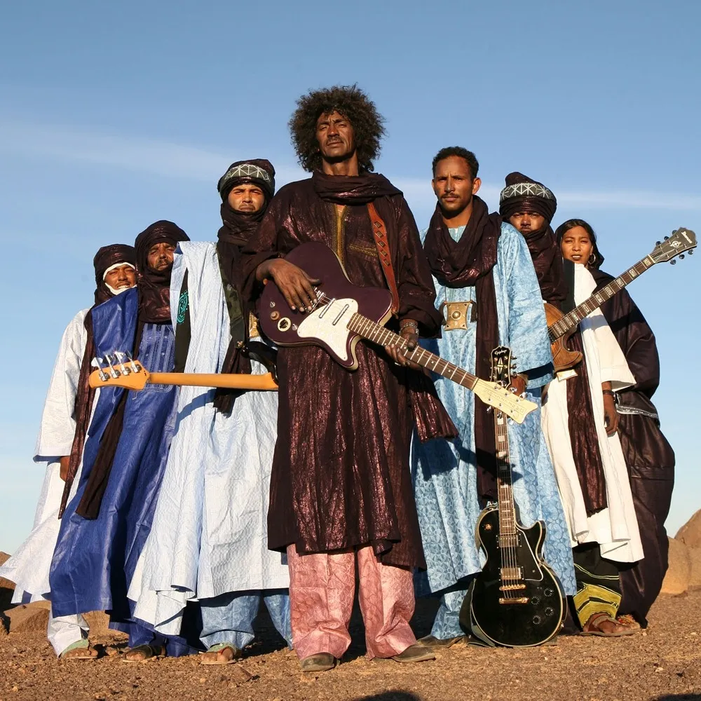 Tinariwen stands outdoors holding electric guitars under a clear blue sky