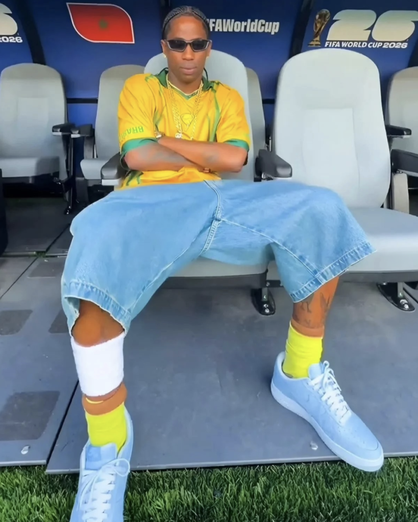 Travis Scott in sunglasses sits in a soccer bench area wearing a yellow Brazil jersey, loose denim shorts, and Travis Scott x Nike Air Force 1 'Ice Blue Ostrich sneakers