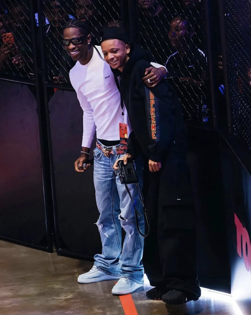 Two smiling young men pose arm-in-arm by a chain-link fence, one holding a camera and wearing black streetwear, the other in a white shirt and jeans