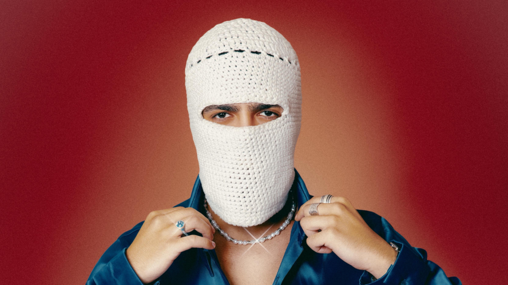 Tul8te in a white crocheted balaclava and blue satin shirt, adjusting the collar against a red background