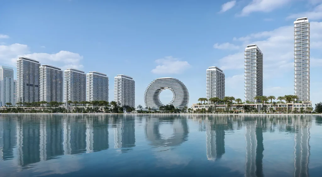 UAQ Marina and Corniche skyline with high-rise towers and a circular ring-shaped building reflected in calm water