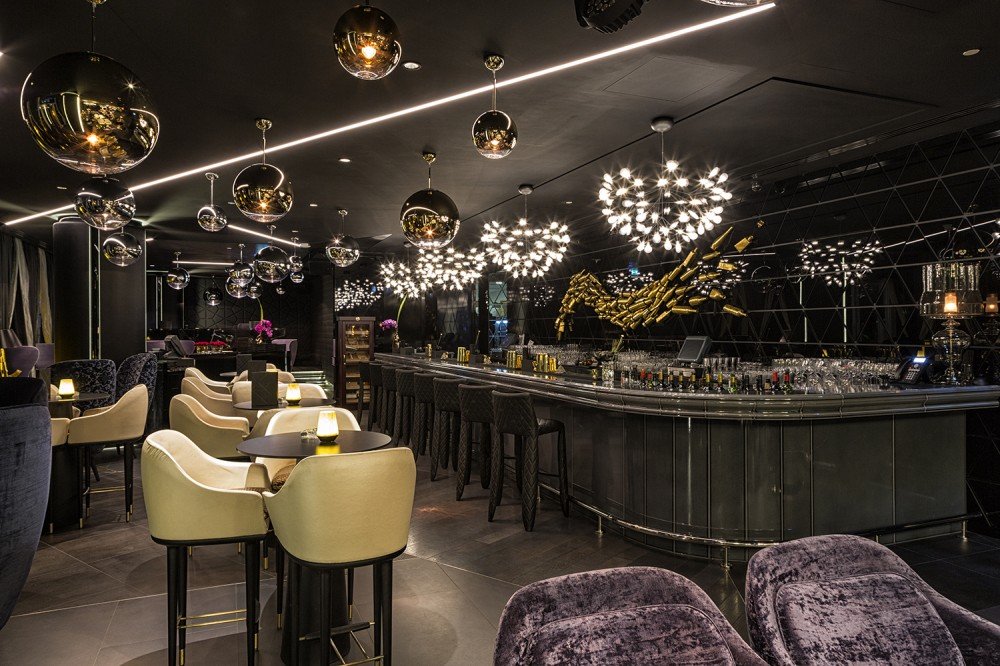 Modern upscale bar lounge with gold accents, pendant lights, tables, chairs, and a long stocked counter