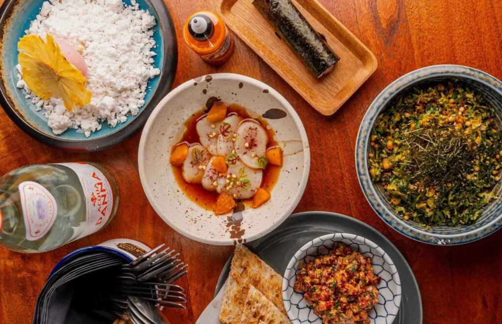 Overhead view of assorted plated dishes and drinks on a wooden table, including seafood, rice, and seaweed soup