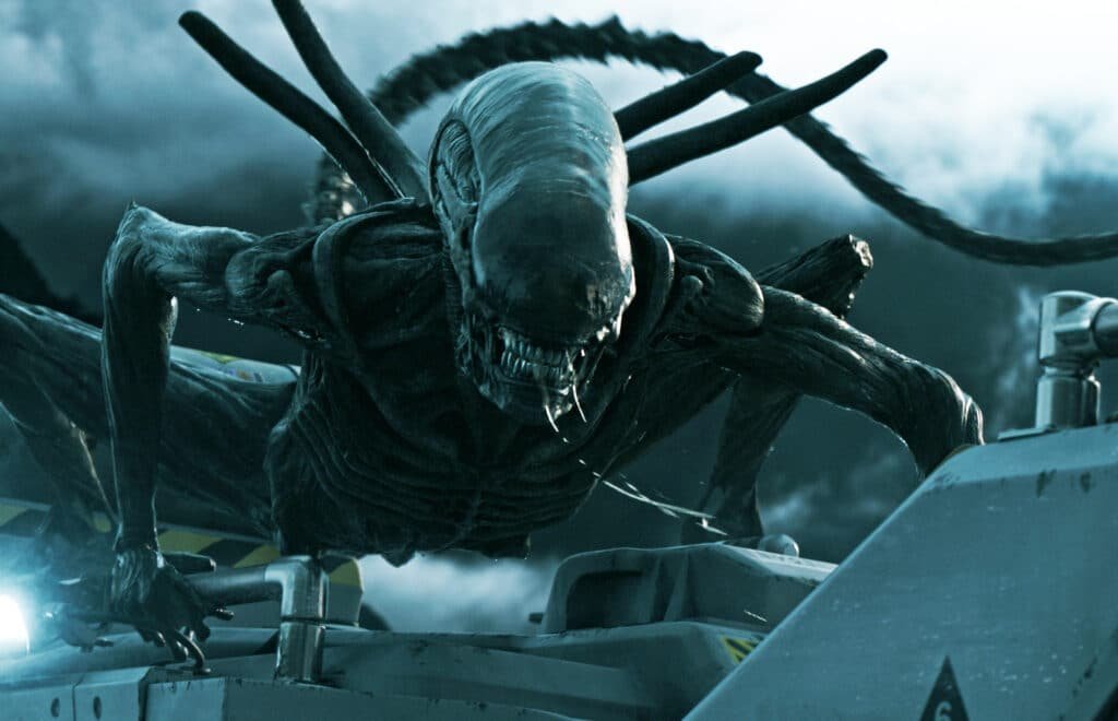 A menacing, toothy Alien prequel creature with elongated limbs crouches on industrial machinery