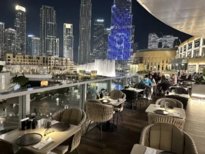 Nighttime rooftop restaurant overlooking Dubai Fountain and illuminated skyscrapers, with diners seated outside